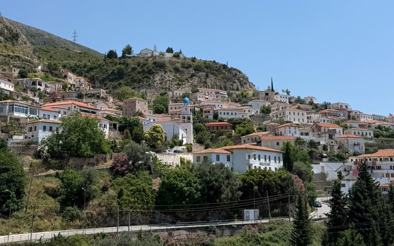 A photograph of Dhërmi, Albania: white houses with terracotta roofs stacked up a green slope around a blue-domed church, with a small chapel alone on the ridge above